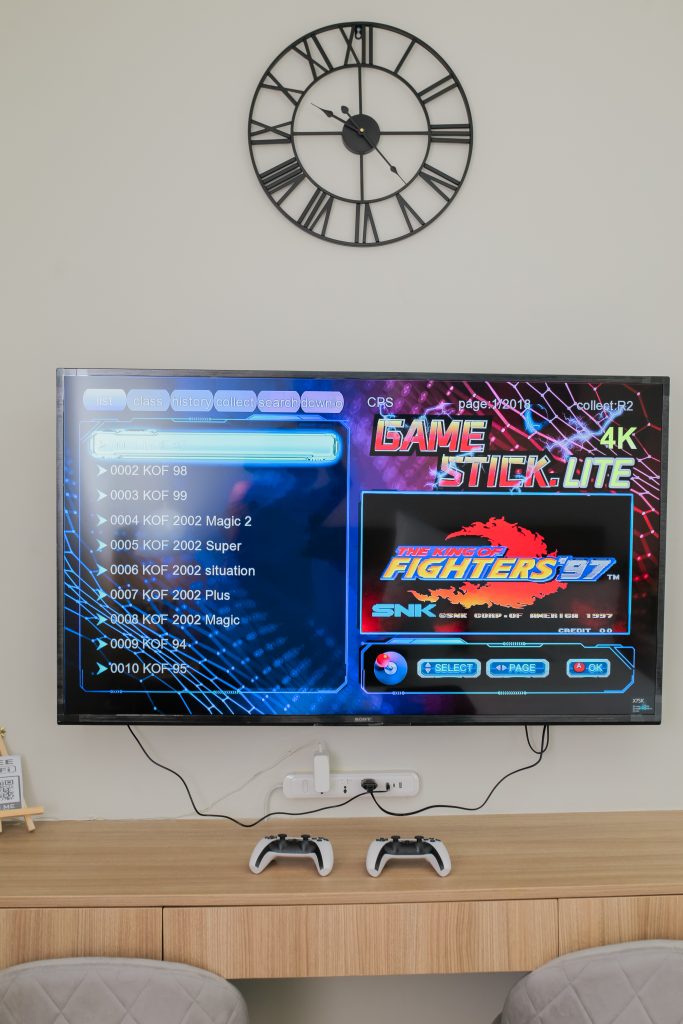 The 55-inch smart TV mounted on the wall displaying the game stick's list of retro games. And a clock above the TV.
